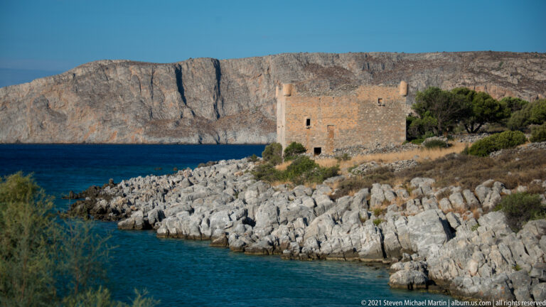 Stone fortified building beside clear turquoise water and angular limestone rocks, with a cliffed headland behind.