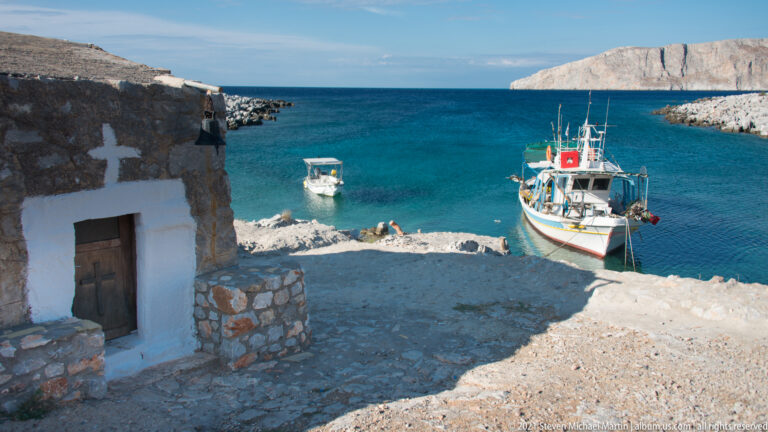 Small whitewashed chapel with a cross beside a blue harbor where two boats are moored.