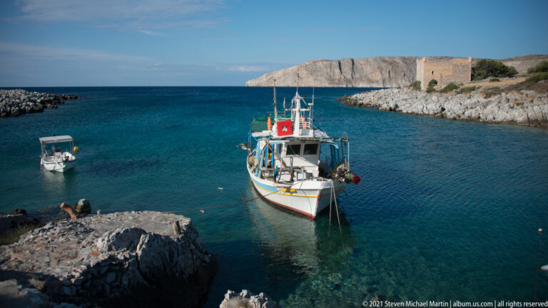 Traditional fishing boat moored in clear blue-green harbor water, with rocky shores and a stone fortification beyond.