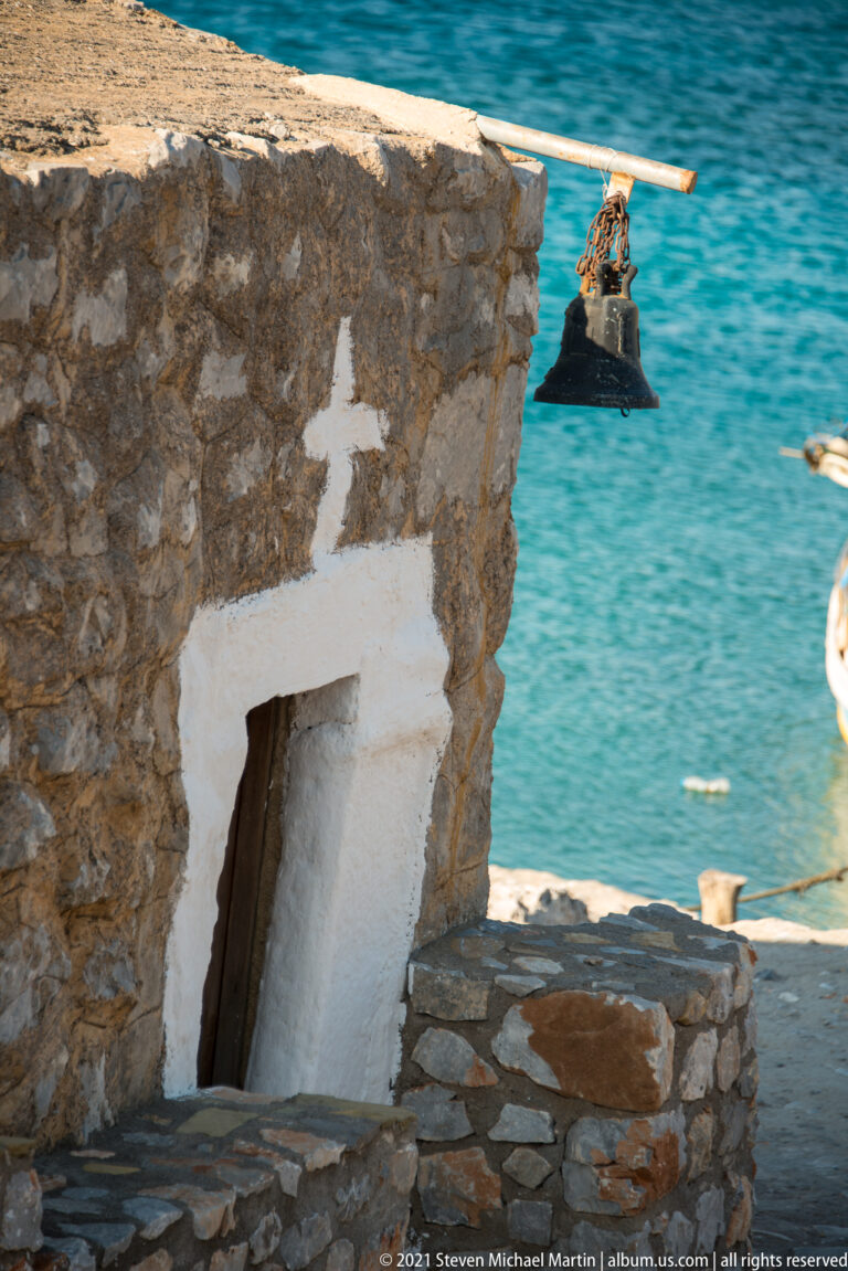 Small stone chapel doorway marked by a white cross, with a hanging bell and turquoise water behind.