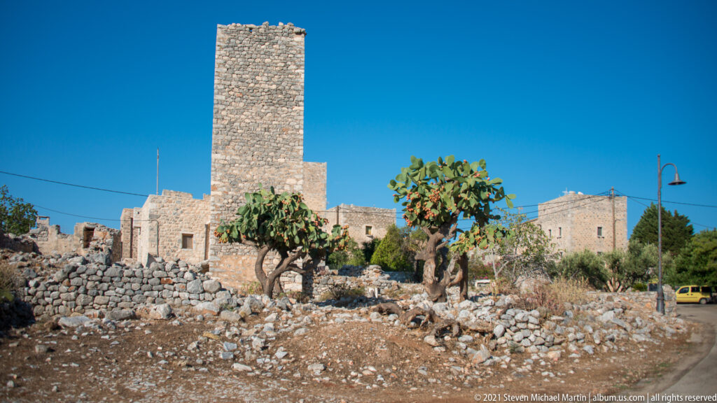 Tall square stone tower above low masonry houses, cactus, and a dry roadside foreground.