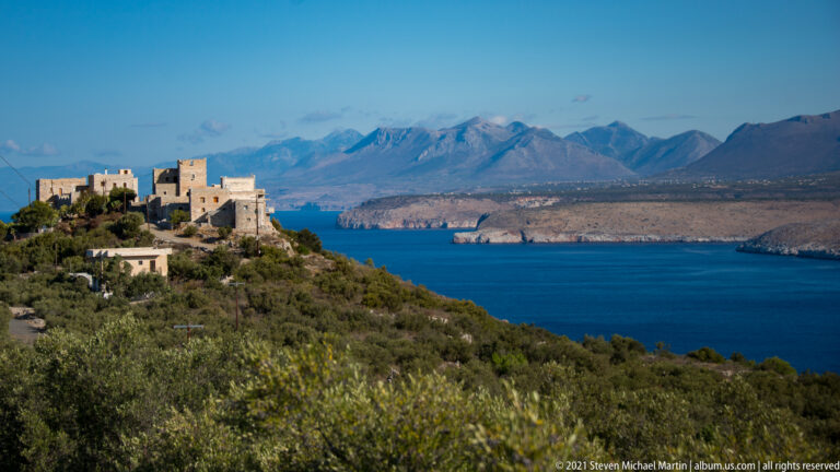 Hilltop cluster of stone houses and tall towers overlooking blue sea and distant mountains.