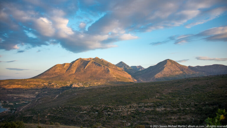 Sunlit rugged mountains rise above dark scrubland, with a small settlement visible near the base of the slopes.