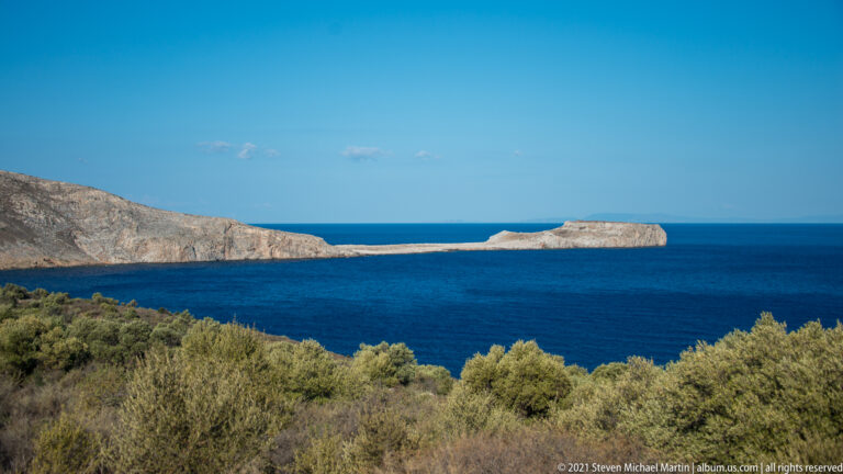 Dry scrub in foreground with pale limestone headlands and a long low offshore rock in deep blue sea.