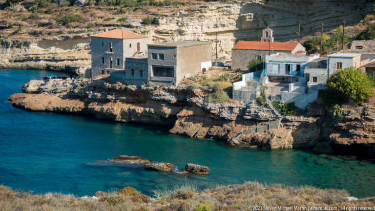 Stone waterfront houses and a small red-roofed church beside a narrow turquoise rocky cove.