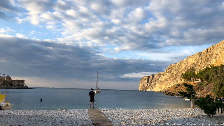 Wooden walkway across a pale pebble beach toward calm water, a sailboat, and high sunlit cliffs.