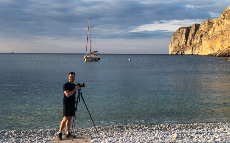 Man standing beside a tripod-mounted camera on a pebble beach, with sailboat and cliffs across the bay.