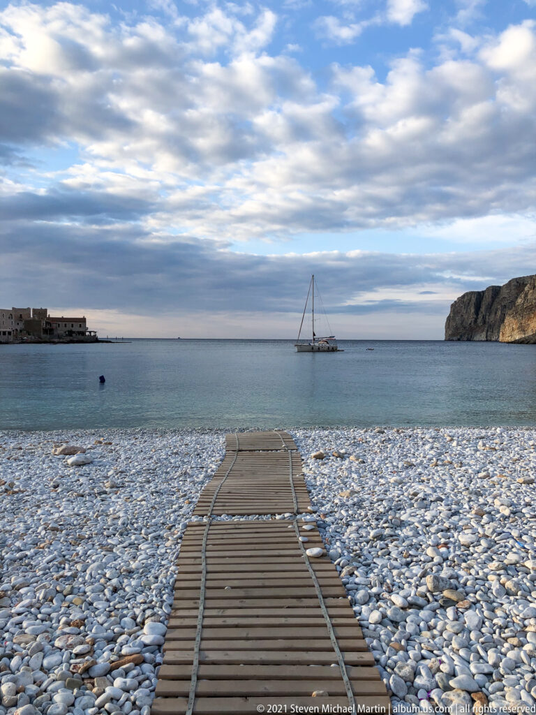 Straight wooden boardwalk crosses a white pebble beach toward calm water and an anchored sailboat.
