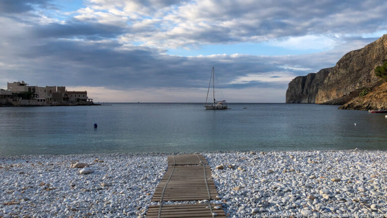 Anchored sailboat in a calm bay between low waterfront buildings and a high limestone cliff, viewed from a pebble beach.
