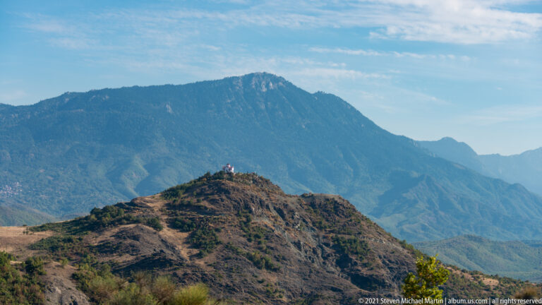 Scrub-covered ridge before a large hazy mountain wall, with a tiny structure on the near crest under blue sky.