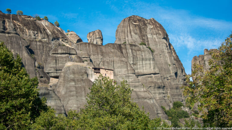 Pale monastery partly hidden between towering gray sandstone walls, with dense trees covering the lower slope.