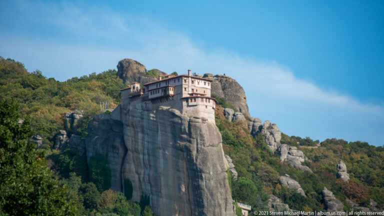 Pale monastery on a tall sandstone pillar viewed across a foreground of dense green trees and shrubs.