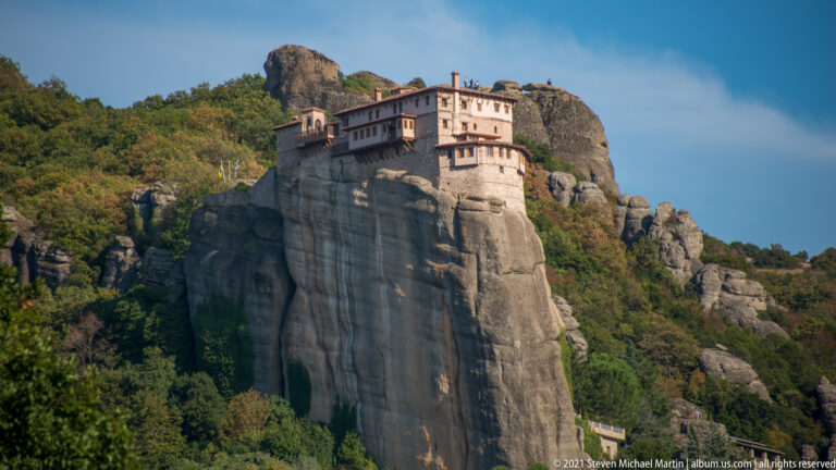 Monastery with pale walls and dark projecting sections atop a sheer gray sandstone pillar above forested slopes.