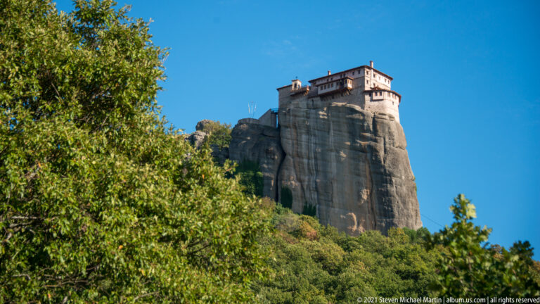 Pale monastery on a narrow sandstone pillar, partly framed by leafy green branches against a blue sky.