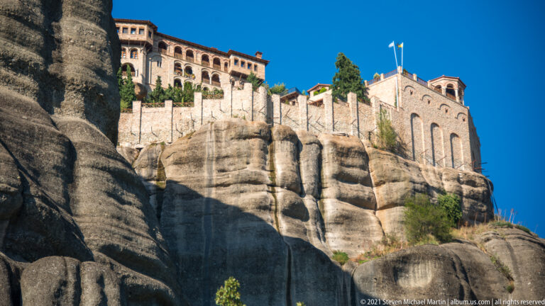 Stone monastery walls with tall arches and upper galleries built directly on rounded sandstone ledges beneath blue sky.