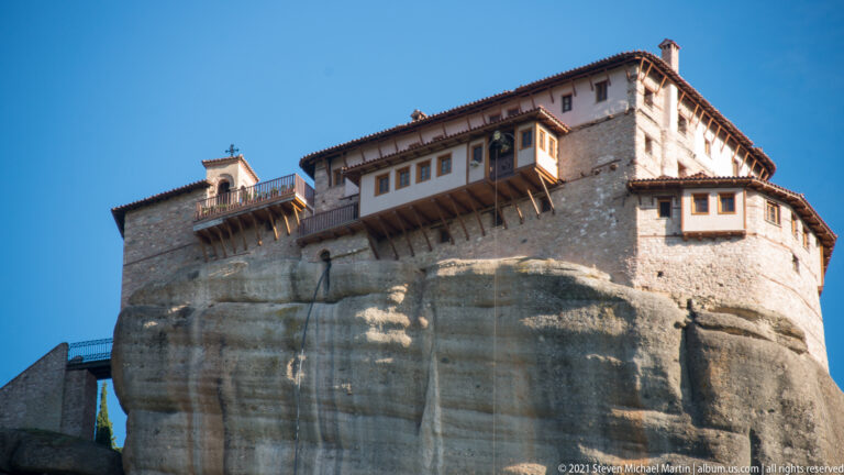 Monastery buildings with projecting timber-supported upper stories perched directly above a rounded sandstone cliff face.