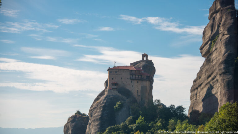 Small monastery on a narrow rock pillar, framed between dark cliffs and backed by a bright, lightly clouded sky.