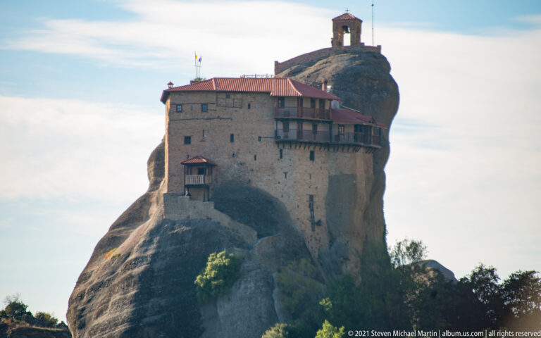 Compact red-roofed monastery atop a narrow sandstone pillar, with a small open tower on a separate rock knob behind.