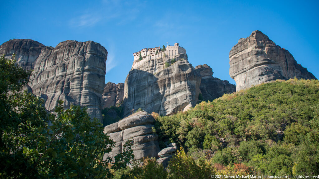 Large pale monastery high among several massive sandstone towers above dense green shrubs and trees.