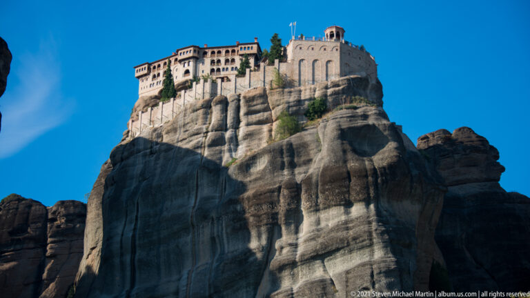 Large sunlit monastery with rows of windows and arched walls perched on a massive rounded sandstone platform.