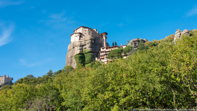 Upper stories of a monastery and adjacent rock towers rising above dense green and yellow foliage.