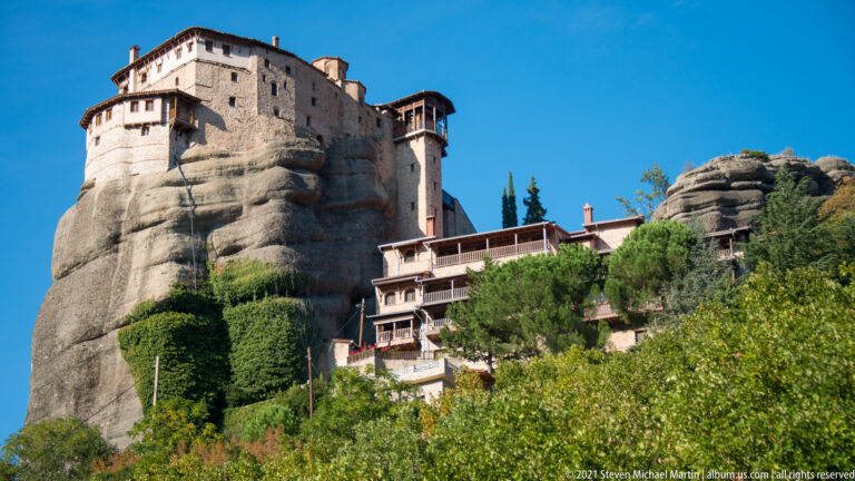 Stone monastery wrapped around a rounded sandstone tower, with terraced buildings descending through trees at right.