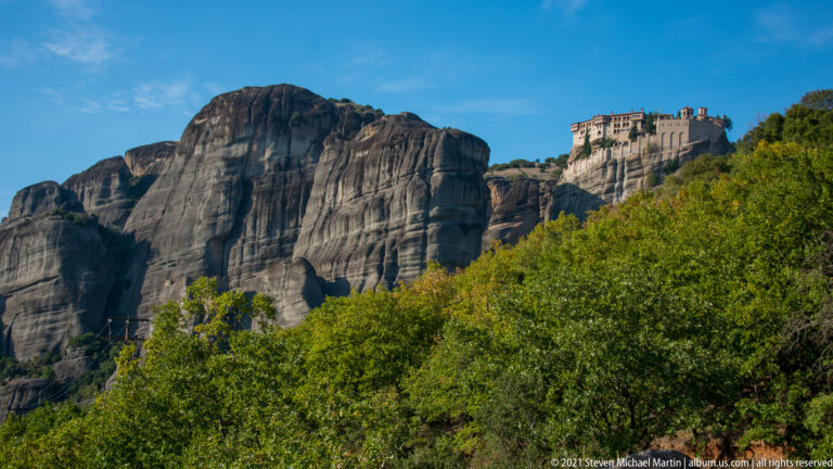 Pale monastery on a distant high rock crest beyond dense foliage, with a massive sandstone wall at left.