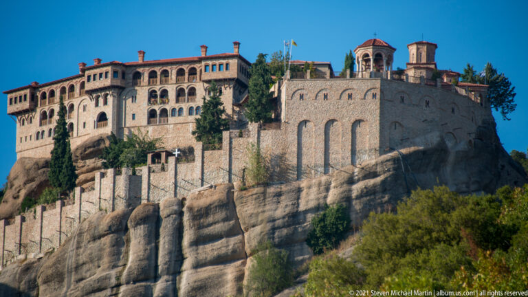 Broad monastery façade with arcaded galleries, stone walls, small towers, and a retaining wall above exposed sandstone.