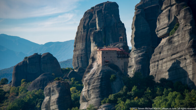 Small red-roofed monastery wedged between enormous dark sandstone pinnacles, with distant mountains visible through a gap.