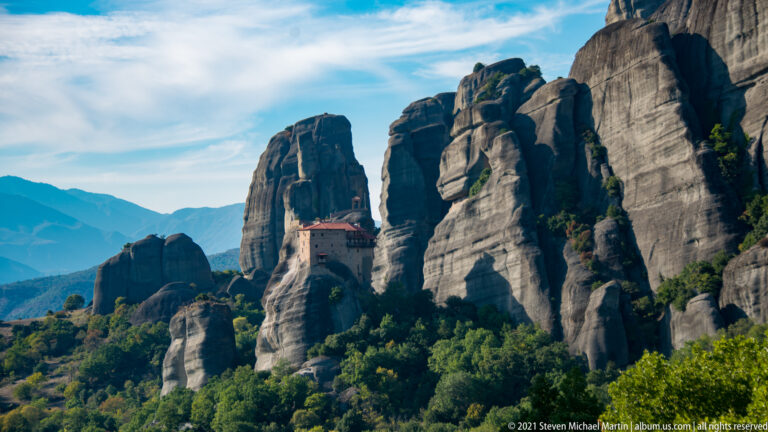 Red-roofed monastery among multiple tall sandstone towers, with forested slopes and distant mountains beyond.