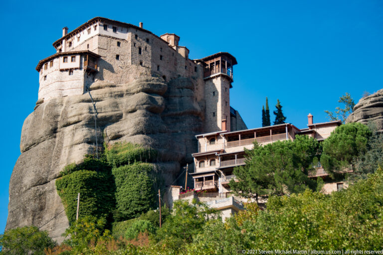 Stone monastery with projecting galleries atop a rounded rock tower, with lower terraces and trees descending at right.