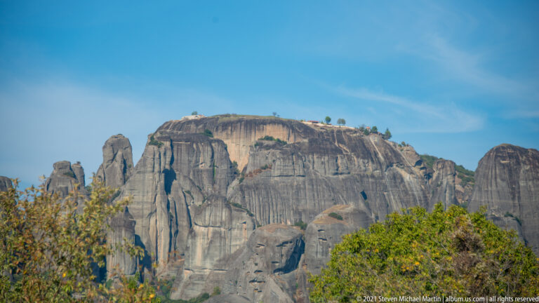 Broad flat-topped sandstone massif with grooved cliffs, sparse trees on the crest, and foliage in the foreground.