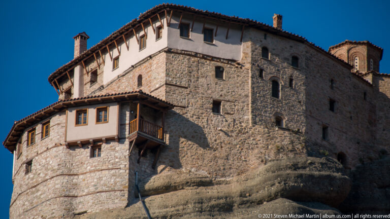 Close view of rough stone and white-plastered monastery walls with tiled roofs, wooden balcony, and rounded rock base.