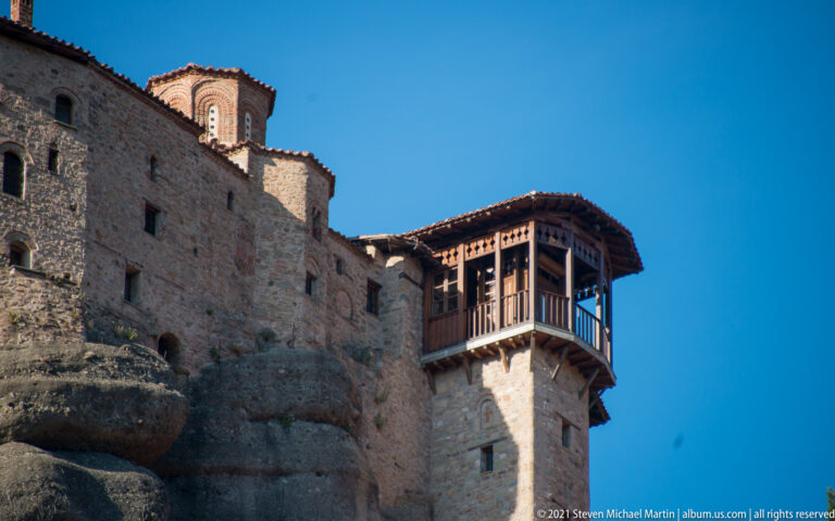 Stone monastery wall built onto exposed rock, with a projecting enclosed wooden gallery beneath a clear blue sky.