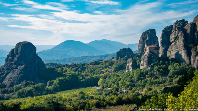 Clustered sandstone towers rise over green forest, with blue mountain ridges layered across the distant horizon.