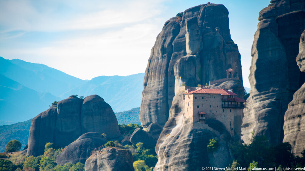 Red-roofed monastery buildings sit atop a narrow sandstone pillar between much taller rock cliffs.