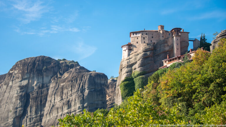 Stone monastery buildings crown a high sandstone outcrop above dense green and autumn-colored trees.