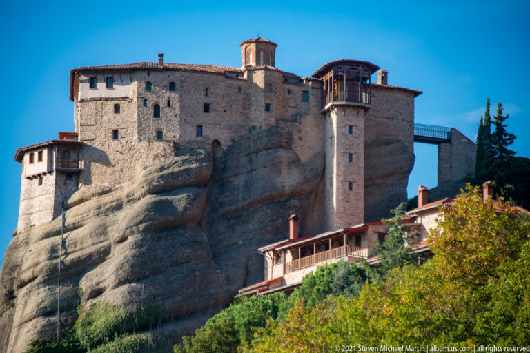 Large stone monastery complex sits on sculpted sandstone, with tiled roofs, a wooden projecting bay, and a narrow bridge.