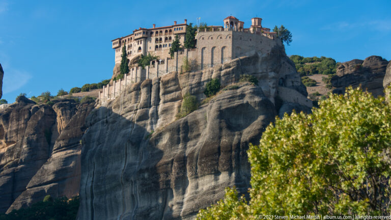 Large monastery complex crowns a high sandstone pillar above steep striated rock faces and trees.
