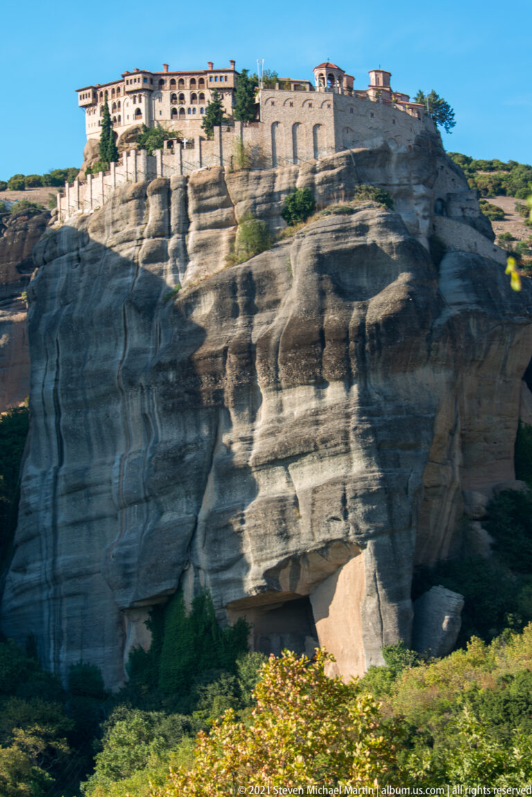 Long monastery buildings line the top of a broad, heavily eroded sandstone cliff with striped rock surfaces.
