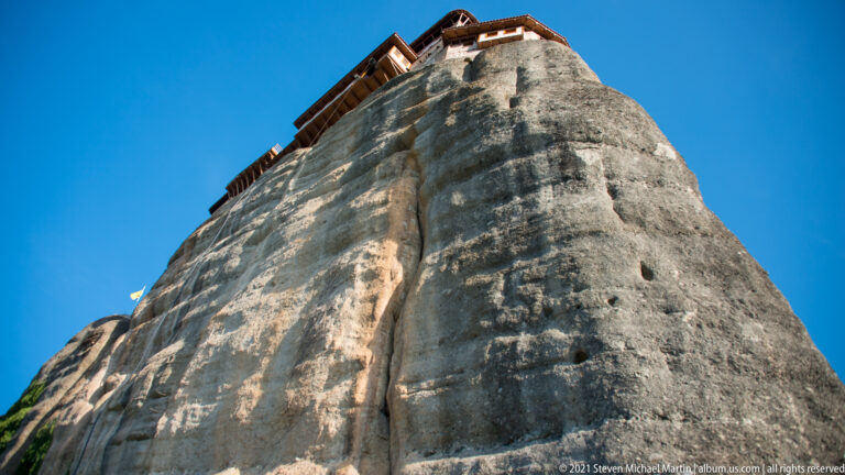 Near-vertical sandstone pillar fills the frame, with monastery rooflines just visible along the summit edge.