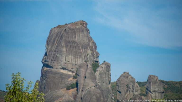 Tall isolated sandstone tower with vertical grooves, smaller pinnacles at its base, and sparse vegetation in cracks.