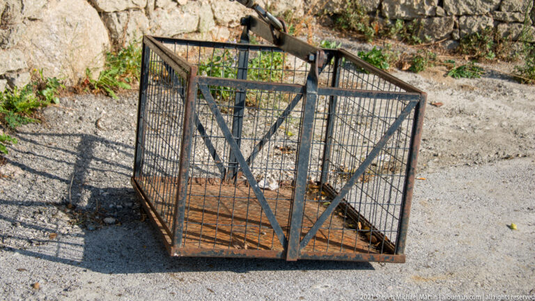 Square iron-bar lifting cage with rusted floor and overhead suspension mechanism standing on gravel beside rock.