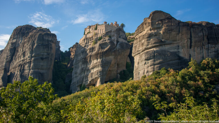 Small distant monastery sits on a rock summit between two massive sandstone towers above dense woodland.