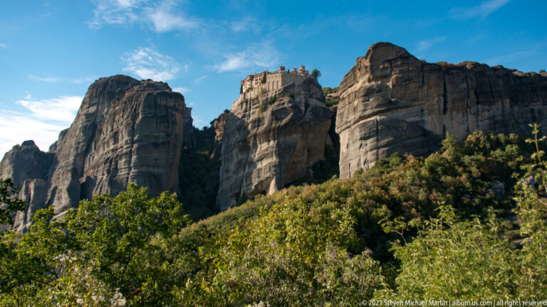 Monastery complex appears between tall sandstone pillars above green wooded slopes in a wide landscape view.