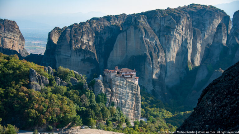 Small red-roofed monastery crowns a narrow rock pillar below much larger sandstone cliffs and a forested ravine.