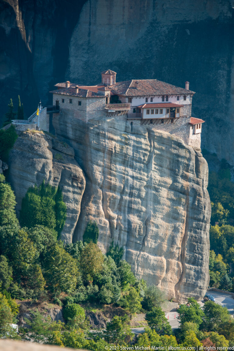 Long monastery complex follows the rounded top of a broad sandstone outcrop above trees and a road.
