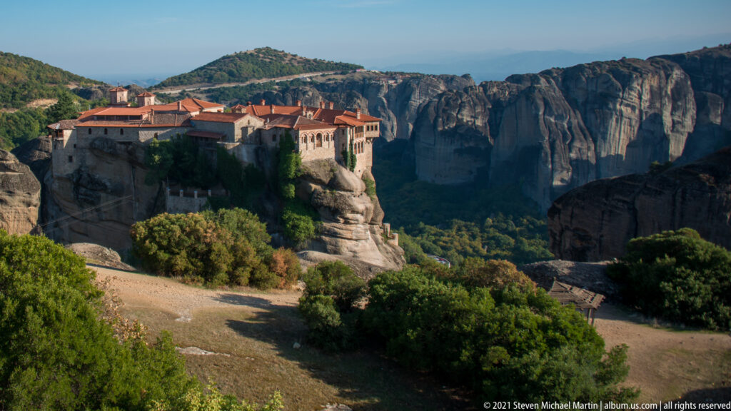 Large red-roofed monastery stands on a high rock platform above a wooded valley and distant cliffs.