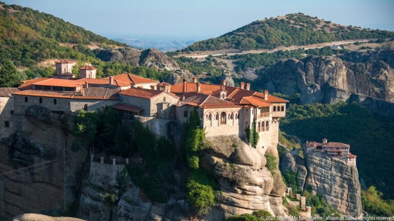 Large foreground monastery and smaller distant monastery sit on separate sandstone pillars amid wooded cliffs.