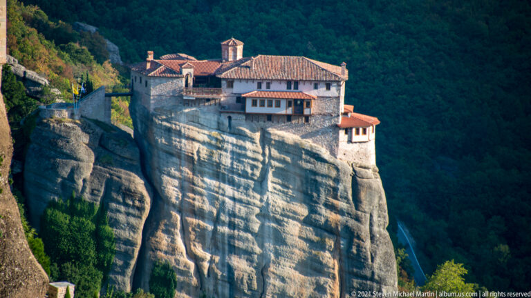 Sunlit stone monastery with red roofs and wooden galleries perches on a sheer sandstone pillar against dark forest.